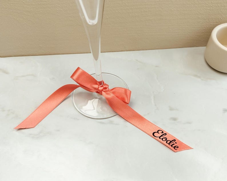 May include: A champagne flute with a coral ribbon tied around the base. The ribbon has the name "Elodie" printed on it in black. The glass is on a white marble surface.