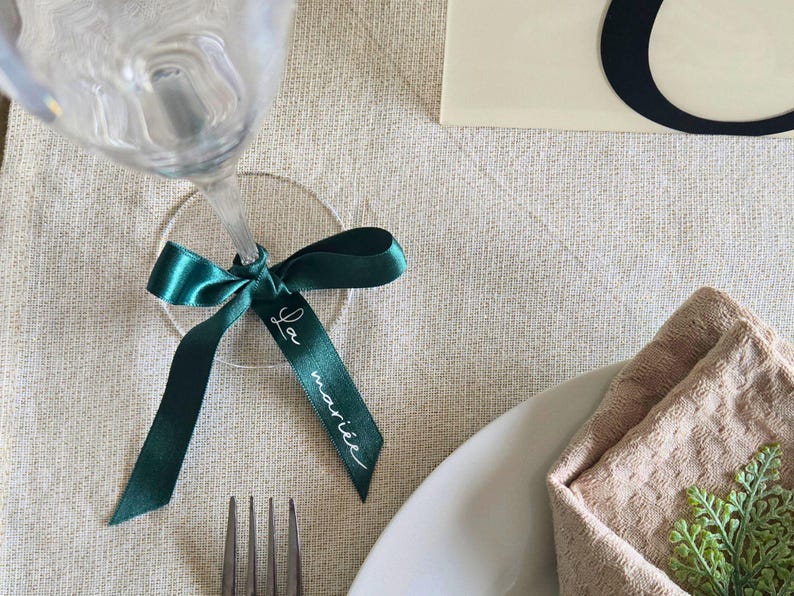 May include: A wine glass with a teal ribbon tied around the stem, featuring the text "La mari&eacute;e" in white script. The setting includes a fork, plate, and napkin, suggesting a wedding or special event.