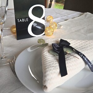 May include: A table setting with a white plate, folded napkin, and a black ribbon with the name "Gabriel". A black and white table number card displays the number "8" and the word "TABLE". Decorative string lights are also present.