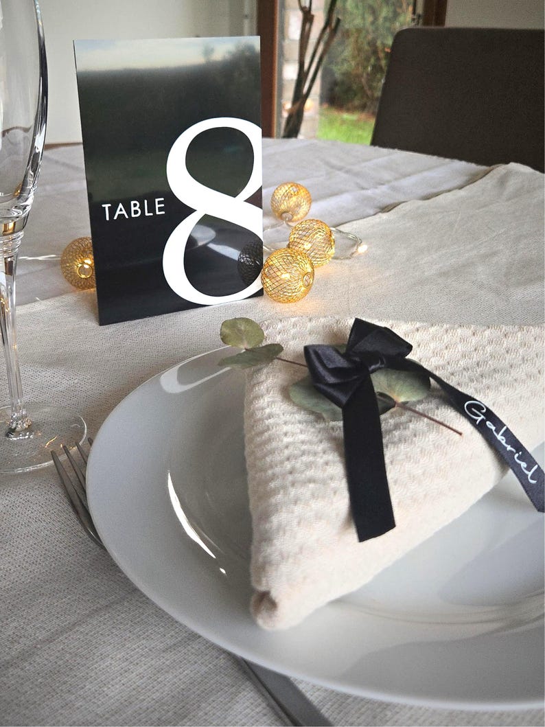 May include: A table setting with a white plate, folded napkin, and a black ribbon with the name "Gabriel". A black and white table number card displays the number "8" and the word "TABLE". Decorative string lights are also present.