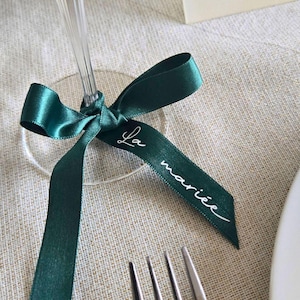 May include: A close-up of a green satin ribbon tied around a clear wine glass. The ribbon has the words "La mari&eacute;e" in white script. The ribbon is on a beige linen tablecloth, with a fork and knife in the foreground.