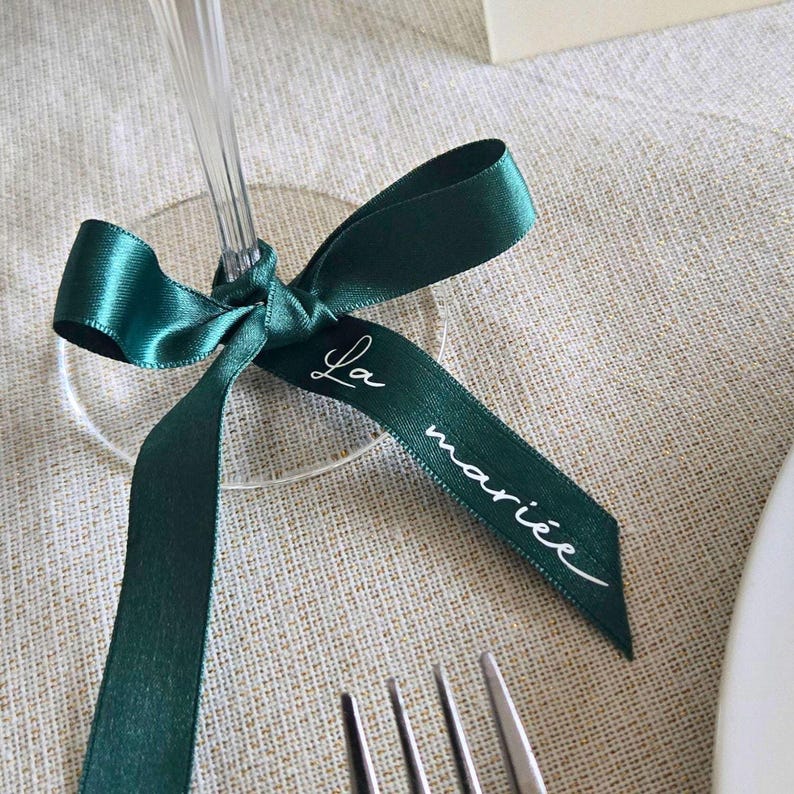 May include: A close-up of a green satin ribbon tied around a clear wine glass. The ribbon has the words "La mari&eacute;e" in white script. The ribbon is on a beige linen tablecloth, with a fork and knife in the foreground.