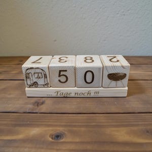 Op de afbeelding: Houten blokkalender met gegraveerde cijfers en afbeeldingen. De blokken tonen de cijfers 7, 3, 8 en 2, samen met een camper en een grill. De basis luidt "... Tage noch !!!". De kalender is gemaakt van lichtgekleurd hout.