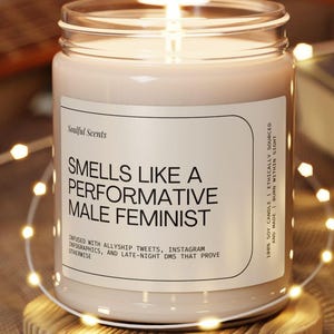 Puede incluir: Una vela encendida en un frasco de vidrio transparente con una etiqueta que dice "Smells Like a Performative Male Feminist". La vela está sobre un pequeño posavasos de madera, con luces de cadena y una guitarra en el fondo.
