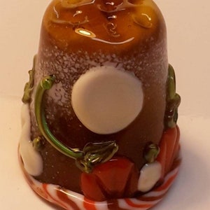 Könnte beinhalten: Ein dekorativer Glasfingerhut mit einem braunen und weißen gesprenkelten Körper, mit einem weißen Kreis und roten, grünen und weißen Akzenten. Der Fingerhut hat ein Kürbisthema mit einem gestreiften Sockel.