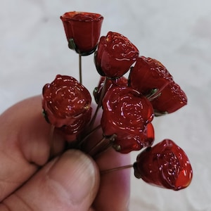 以下が含まれることがあります： 緑色のアクセントが付いた、ガラスのような小さな赤いバラの束。 バラは細い茶色の茎にまとめられています。 花は光沢のある仕上がりで、手で持たれています。