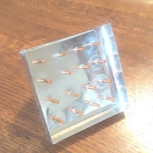 May include: A clear acrylic display stand with a grid of 20 small, copper-colored metal rods. The stand is angled and has a reflective surface.