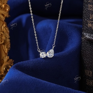 May include: A delicate silver necklace with two clear, sparkling gemstones. One gemstone is round, and the other is a pear shape. The necklace is displayed on a deep blue fabric, creating a contrast that highlights the jewelry.