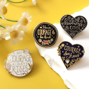 Könnte beinhalten: Vier Emaille-Anstecknadeln mit inspirierenden Zitaten. Eine runde Nadel sagt "Have COURAGE & be kind". Zwei herzförmige Nadeln lauten "kindness is COOL" und "COURAGE dear heart". Eine vierte Nadel sagt "MY COURAGE ALWAYS RISES."