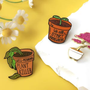 Może przedstawiać: Trzy emaliowane przypinki na żółtym i białym tle. Jedna przypinka to drzewko bonsai z różowym, błyszczącym wierzchołkiem. Pozostałe dwie przypinki to pomarańczowe doniczki z zielonymi roślinami i tekstem "We all START SOMEWHERE" i "WELL-MEANING PLANT KILLER."