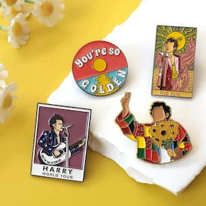 Può includere: Una collezione di spille smaltate con illustrazioni di un cantante. Le spille includono un design circolare con la scritta "you're so golden", una spilla rettangolare con la scritta "HARRY WORLD TOUR" e altre due spille con l'immagine del cantante.
