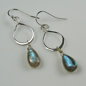 May include: A pair of silver-toned earrings with a teardrop shape. Each earring features a silver infinity loop with a teardrop-shaped labradorite gemstone pendant. The gemstones have a mix of gray, blue, and green hues.
