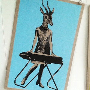 Op de afbeelding: Een ingelijste kunstprint met een gestileerde figuur met een antilopenkop die een keyboard bespeelt. De figuur draagt een jurk en laarzen, tegen een lichtblauwe achtergrond. De print wordt vastgehouden met metalen clips.