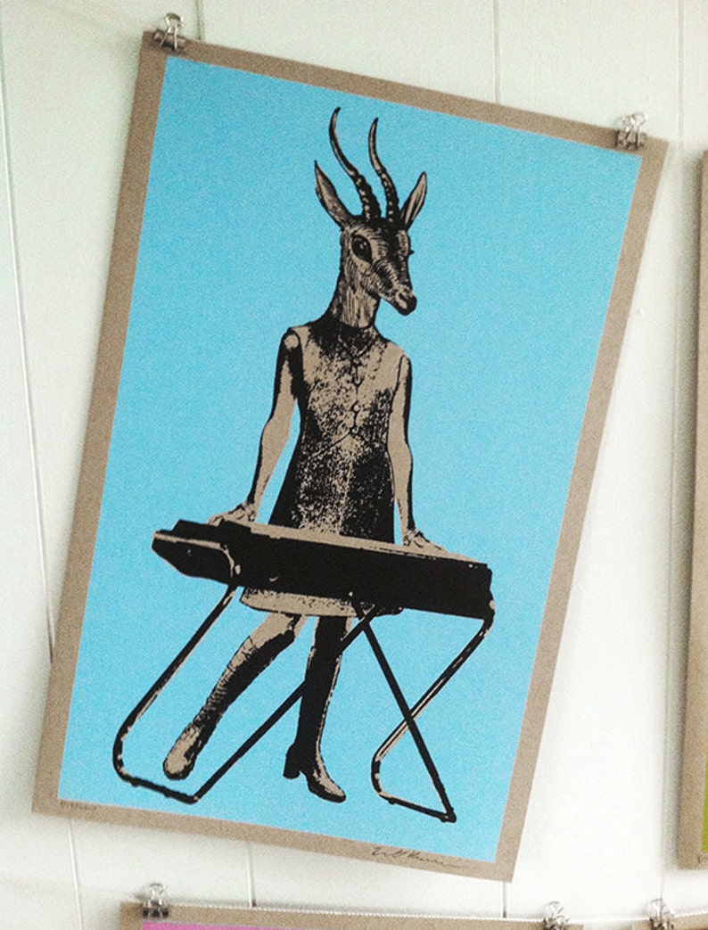 Op de afbeelding: Een ingelijste kunstprint met een gestileerde figuur met een antilopenkop die een keyboard bespeelt. De figuur draagt een jurk en laarzen, tegen een lichtblauwe achtergrond. De print wordt vastgehouden met metalen clips.