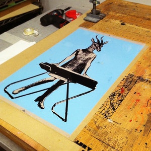 Op de afbeelding: Een zeefdruk op lichtblauw papier met een figuur met een hertenkop die een keyboard bespeelt. De figuur is zwart-wit. De print staat op een houten tafel met de tekst "LERY 001 SHOW" in de onderste hoek.