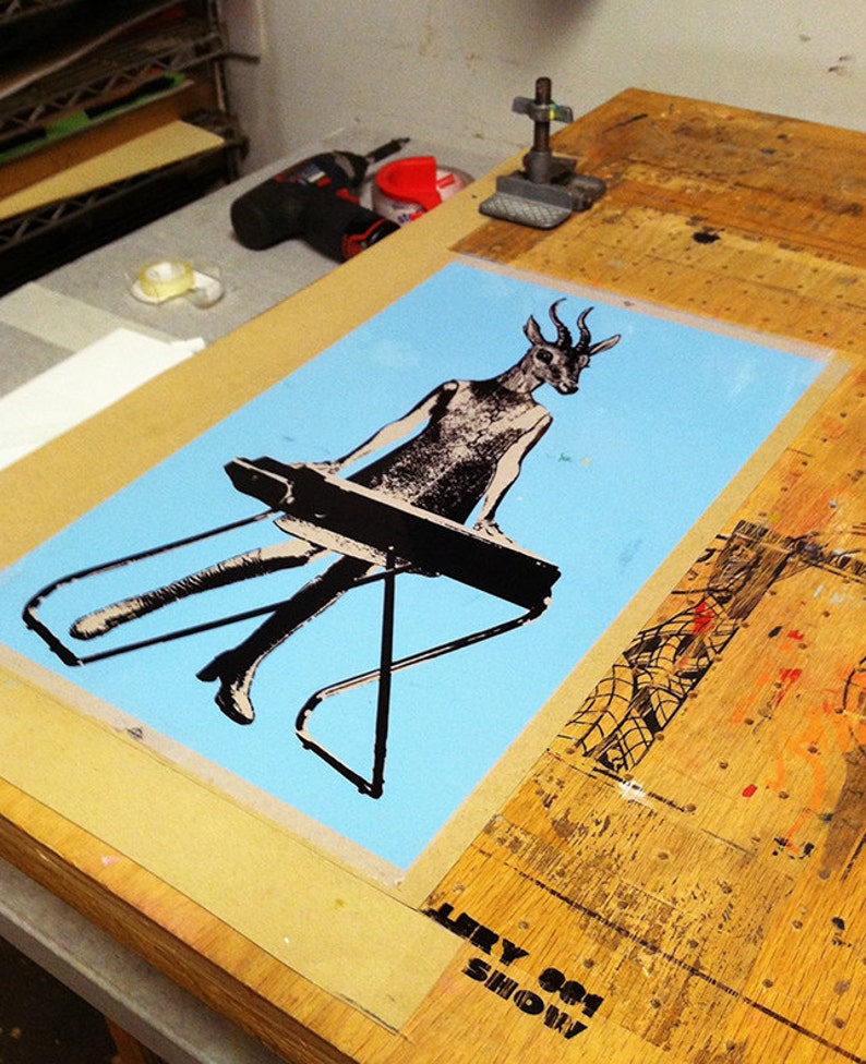 Op de afbeelding: Een zeefdruk op lichtblauw papier met een figuur met een hertenkop die een keyboard bespeelt. De figuur is zwart-wit. De print staat op een houten tafel met de tekst "LERY 001 SHOW" in de onderste hoek.