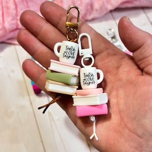 May include: Two keychains with a gold and white clasp. Each keychain features a white mug with the text "Teach Love Inspire" and a stack of books in various colors. The background is a light pink.