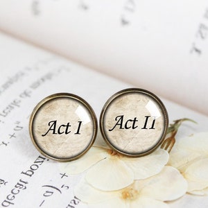 Op de afbeelding: Een paar ronde, bronzen manchetknopen met glazen inzetstukken. De glazen inzetstukken zijn beige met een vintage papiertextuur en zwarte tekst. De tekst op de linker manchetknoop is "Act 1" en de tekst op de rechter manchetknoop is "Act 11".