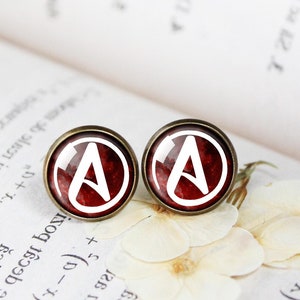 Może przedstawiać: Para kolczyków z czerwonym tłem i białym symbolem anarchii. Kolczyki są wykonane z metalu i mają okrągły kształt.