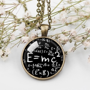 Può includere: Una collana bianca e nera con un ciondolo rotondo che presenta la famosa equazione E=mc2 e altre formule scientifiche. Il ciondolo è circondato da una cornice in metallo color bronzo.