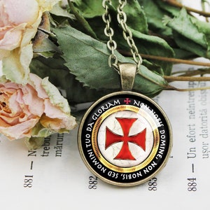 May include: A silver chain necklace with a round glass pendant featuring a red cross on a white background. The pendant has a gold rim and the text "NON NOBIS, SED NOBIS DOMINE, TUO DA GLORIAM" in black lettering.