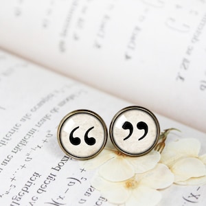 May include: A pair of small, round, cream-coloured earrings with a bronze rim. Each earring features a black graphic of quotation marks.