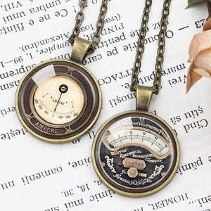 Peut inclure: Deux colliers à chaîne dorée avec des pendentifs ronds en verre représentant des jauges analogiques de style vintage. Les jauges sont marquées en noir et blanc et le texte "AMPERE" et "WESTON VOLTMETER" est visible.