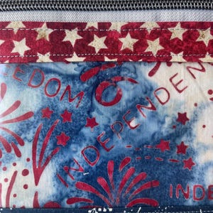 May include: Close-up of a fabric item with a zipper and patriotic design. The design features red stars on a white background, and a blue background with red fireworks and the word "INDEPENDENCE".