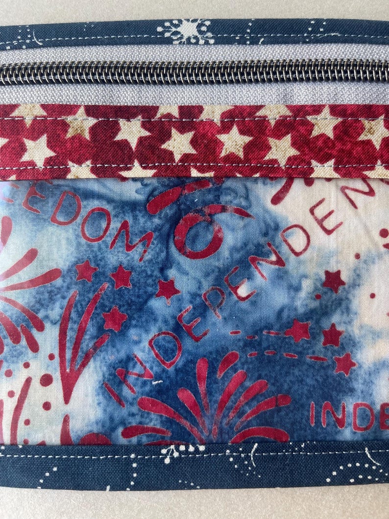 May include: Close-up of a fabric item with a zipper and patriotic design. The design features red stars on a white background, and a blue background with red fireworks and the word "INDEPENDENCE".
