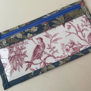 May include: A rectangular pencil case with a clear window. The case features a blue zipper and is trimmed with a floral fabric in shades of blue, green, and cream. The window displays a red and white bird and floral pattern.