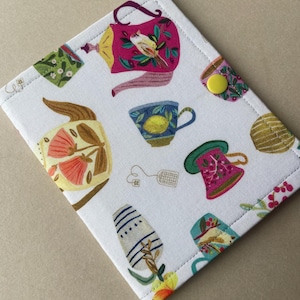 May include: A white fabric wallet featuring a colorful pattern of teapots, teacups, and tea bags in shades of pink, yellow, blue, and green. The wallet is secured with a single bright yellow snap button and has visible stitching along the edges.