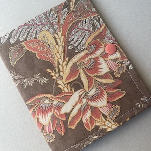 May include: A rectangular fabric cover featuring a brown background with a repeating floral pattern in shades of cream, peach, and dark red. The cover is secured with a single, round, solid peach-colored plastic snap button on the right side.