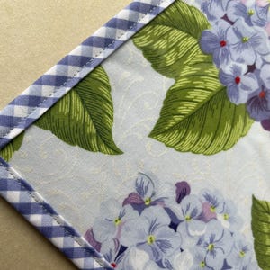 May include: Close-up of a floral fabric with a blue and white gingham border. The fabric features a pattern of blue, purple, and white hydrangea flowers with green leaves. The background has a subtle, light-colored pattern.