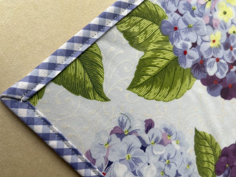 May include: Close-up of a floral fabric with a blue and white gingham border. The fabric features a pattern of blue, purple, and white hydrangea flowers with green leaves. The background has a subtle, light-colored pattern.