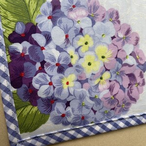 May include: A floral fabric with a hydrangea design in shades of purple, lavender, and yellow. The fabric is bordered with a blue and white gingham pattern. Green leaves are visible.