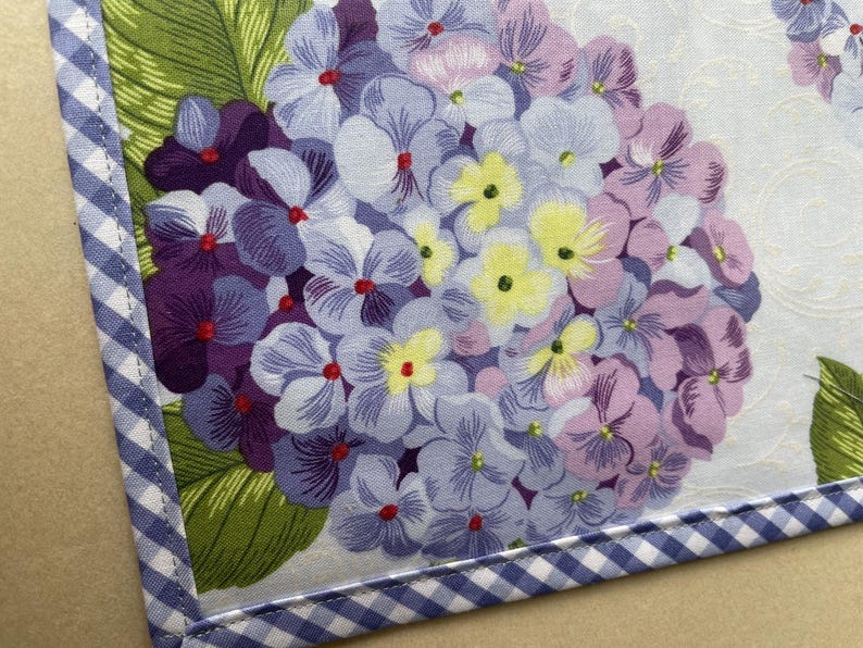 May include: A floral fabric with a hydrangea design in shades of purple, lavender, and yellow. The fabric is bordered with a blue and white gingham pattern. Green leaves are visible.
