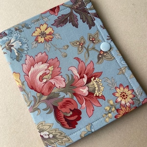 May include: A rectangular fabric case featuring a light blue background with a floral pattern in shades of red, pink, yellow, and muted green. The case is secured with a single light blue plastic snap button and has visible stitching along the edges.