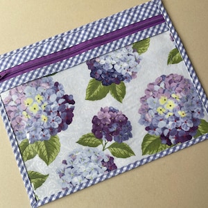 May include: A rectangular zippered pouch featuring a floral hydrangea pattern in shades of purple, blue, and yellow on a white background. The pouch is trimmed with a blue and white gingham pattern and a purple zipper.