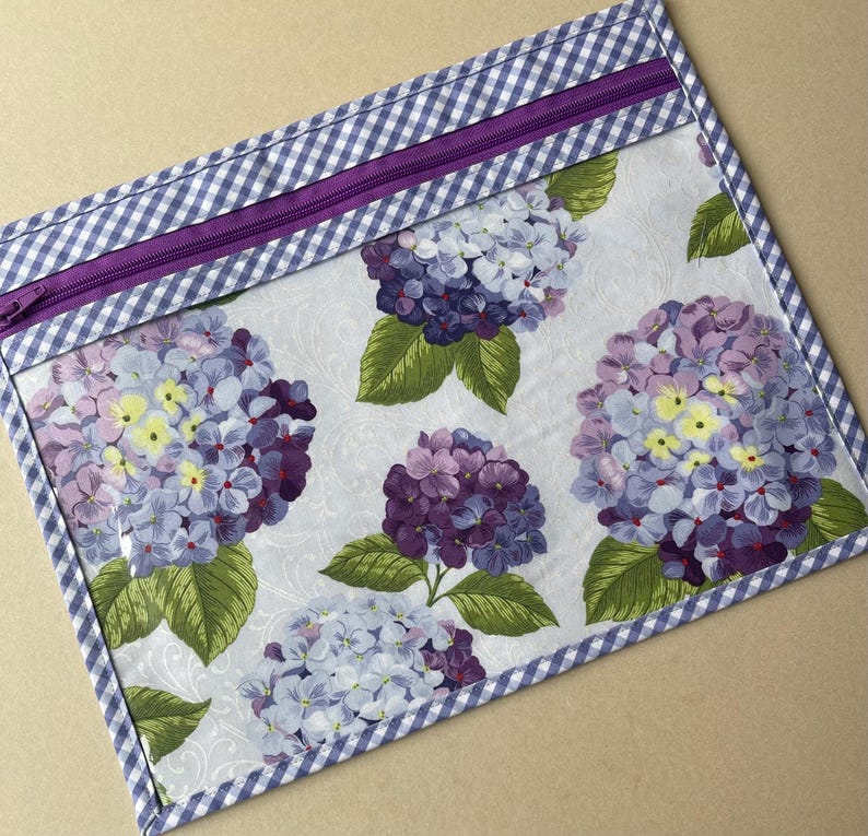 May include: A rectangular zippered pouch featuring a floral hydrangea pattern in shades of purple, blue, and yellow on a white background. The pouch is trimmed with a blue and white gingham pattern and a purple zipper.