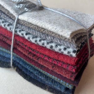 May include: A stack of rectangular fabric swatches tied together with thin gray string. The collection includes layers of felted wool in various colors and textures, featuring solid light gray, a black and white speckled pattern, deep red, and dark blue.