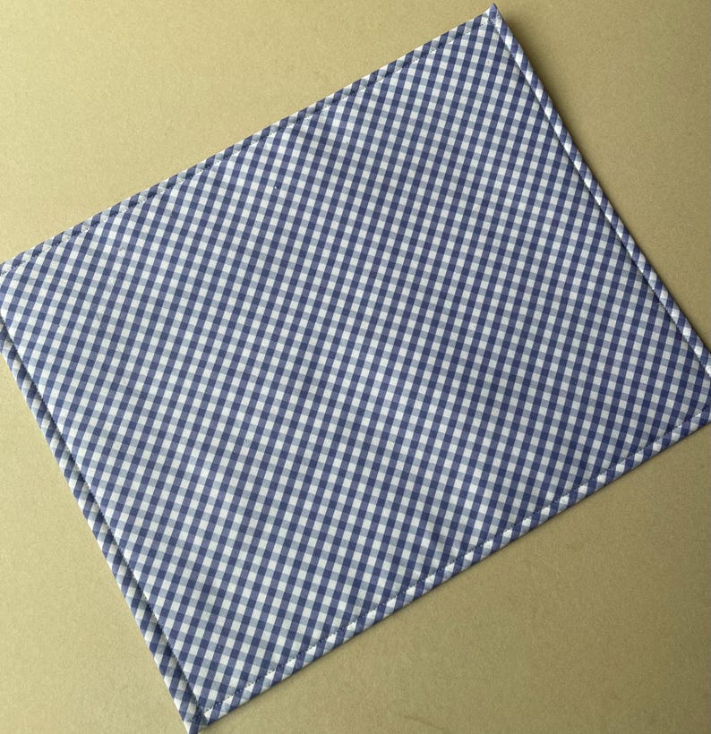 May include: A blue and white checkered placemat. The placemat has a classic gingham pattern, with a stitched edge. The fabric is a light blue and white, creating a simple, yet elegant design. The placemat is rectangular.