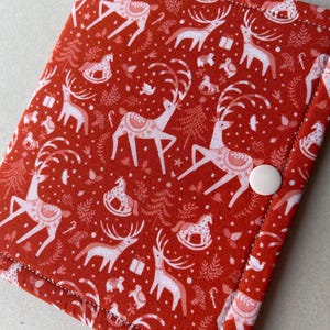 May include: Red fabric item with a white Christmas pattern featuring reindeer, rocking horses, and gifts. The fabric is secured with white stitching and a white button closure. The item is likely a small case or wallet.