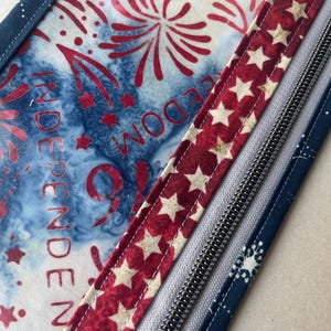 May include: Close-up of a patriotic-themed fabric with red, white, and blue colors. The design includes fireworks, stars, and the word "INDEPENDENCE" and "FREEDOM". A red ribbon with white stars and a silver zipper are also visible.