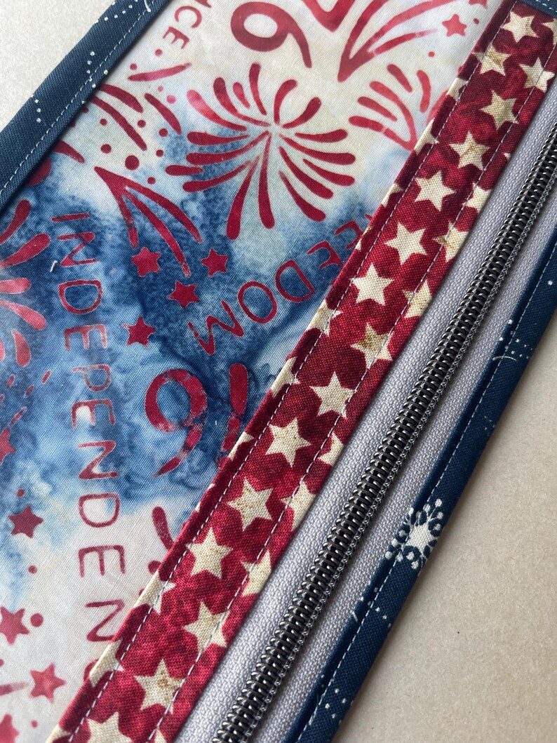May include: Close-up of a patriotic-themed fabric with red, white, and blue colors. The design includes fireworks, stars, and the word "INDEPENDENCE" and "FREEDOM". A red ribbon with white stars and a silver zipper are also visible.