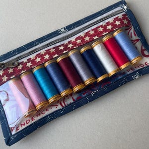 May include: A rectangular, zippered sewing kit with a patriotic theme. The kit features a navy blue fabric with white star and dot patterns, a red and white star-patterned accent, and a clear window displaying spools of colorful thread.
