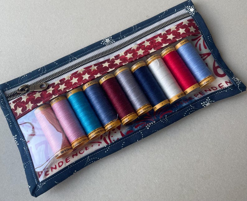 May include: A rectangular, zippered sewing kit with a patriotic theme. The kit features a navy blue fabric with white star and dot patterns, a red and white star-patterned accent, and a clear window displaying spools of colorful thread.