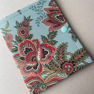 May include: A rectangular fabric cover featuring a light blue background with a vibrant floral pattern in shades of pink, red, dark green, and cream. The cover is secured with a single light blue snap button located on the right side edge.
