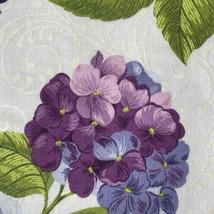 May include: Close-up of a fabric with a floral pattern. The design features a cluster of purple and lavender hydrangea flowers with green leaves. The background is a light cream color with a subtle, swirling pattern.
