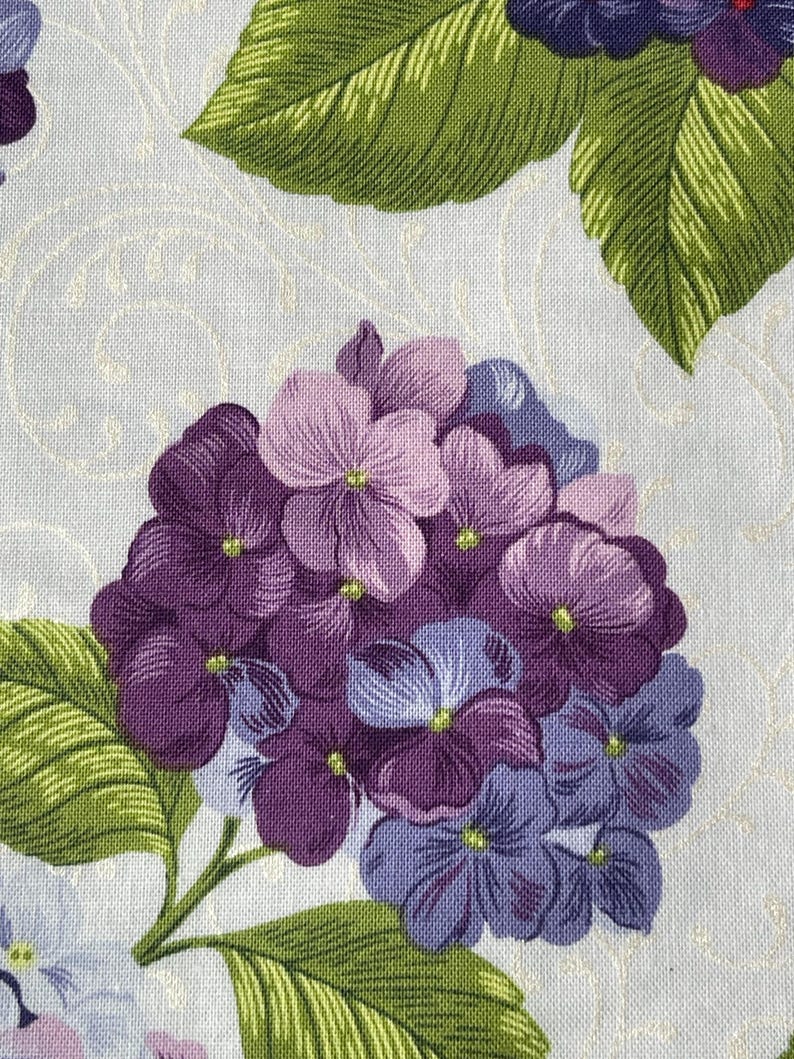 May include: Close-up of a fabric with a floral pattern. The design features a cluster of purple and lavender hydrangea flowers with green leaves. The background is a light cream color with a subtle, swirling pattern.