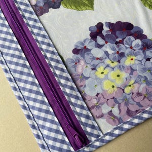 May include: A close-up of a fabric item featuring a purple zipper and a blue and white gingham pattern. The main fabric displays a floral design with purple, blue, and yellow hydrangea blooms and green leaves.